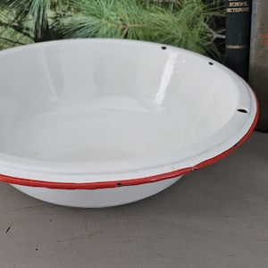 May include: A white enamelware bowl with a red rim. The bowl is round and has a wide, shallow design. It has a few small holes near the rim. The bowl is sitting on a gray surface.