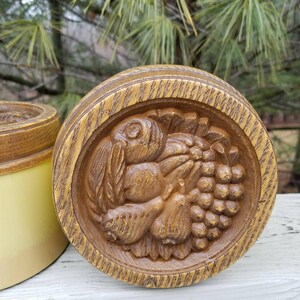 May include: A round, brown, wood-toned container lid featuring a detailed relief carving of a cluster of grapes, a pear, and leaves. The circular lid has a textured, grain-like finish and sits beside a smooth, light yellow cylindrical container base.