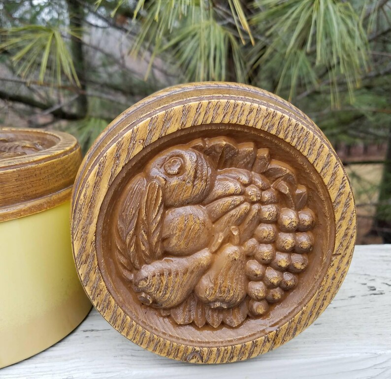 May include: A round, brown, wood-toned container lid featuring a detailed relief carving of a cluster of grapes, a pear, and leaves. The circular lid has a textured, grain-like finish and sits beside a smooth, light yellow cylindrical container base.