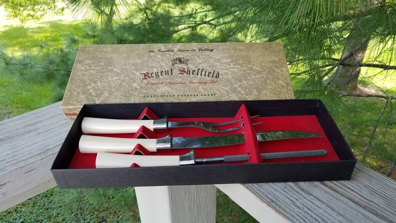 Puede incluir: Un juego de cuatro cuchillos de plata con mangos de marfil en una caja negra con forro de terciopelo rojo. La tapa de la caja est&aacute; abierta y presenta el texto "Regent Sheffield English Stainless Cutlery Set Guaranteed Forever Sharp".
