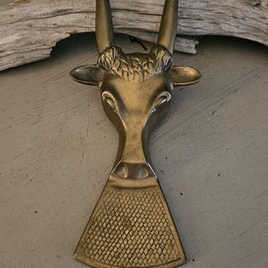 May include: A brass bottle opener in the shape of a deer head. The opener features curved antlers, a detailed face, and a textured, diamond-patterned base. The opener is a decorative and functional item.