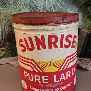 May include: Vintage Sunrise Pure Lard tin with a red lid and accents. The cream-colored can features red lettering and a sunburst design. It reads "Sunrise Pure Lard" and "American Packing Company" and weighs 25 pounds.
