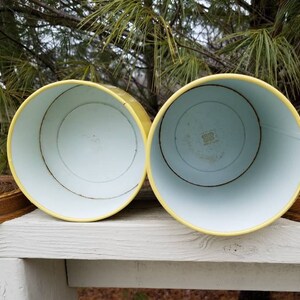 May include: Two cylindrical metal canisters with bright yellow exteriors and white interiors. Each canister is accompanied by a separate, circular, brown textured lid featuring a detailed relief pattern on the top surface.