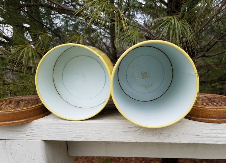 May include: Two cylindrical metal canisters with bright yellow exteriors and white interiors. Each canister is accompanied by a separate, circular, brown textured lid featuring a detailed relief pattern on the top surface.