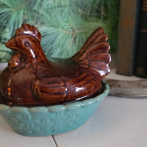 May include: A brown ceramic chicken figurine sitting in a teal blue ceramic nest. The chicken has a glossy finish and is detailed with brown and white markings.