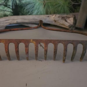 May include: An antique garden rake with a rusty, weathered appearance. The rake features a series of curved tines and a curved handle, all showing signs of age and use. The metal has a mix of orange, brown, and green hues.