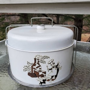 May include: White metal cake carrier with a brown and black illustration of a chef and a fireplace. The carrier has a wire handle and a removable lid.