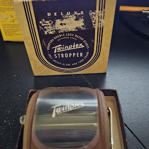 May include: A vintage Twinplex razor blade stropper with a brown casing and a clear top, housed in its original cardboard box. The box features the text 'DELUXE', 'Twinplex STROPPER', and 'SHARPENS DOUBLE EDGE RAZOR BLADES (GILLETTE TYPE)'.