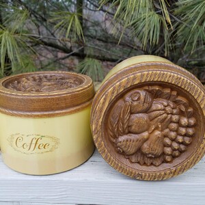 May include: A cylindrical cream-colored coffee canister with a brown wood-grain textured lid. The lid features a detailed relief carving of assorted fruits including grapes and pears. The word Coffee is printed in brown script on the front side.