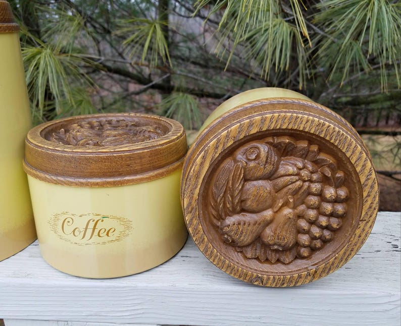 May include: A cylindrical cream-colored coffee canister with a brown wood-grain textured lid. The lid features a detailed relief carving of assorted fruits including grapes and pears. The word Coffee is printed in brown script on the front side.
