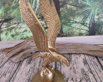 May include: A gold-colored metal eagle sculpture with outstretched wings, perched atop a globe and a circular base. The eagle's feathers are intricately detailed. The base has engraved text. The sculpture is displayed on a wooden surface.