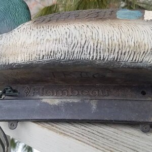 May include: A green and brown duck decoy with a black base. The decoy is made by Flambeau and has a realistic design.