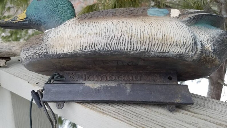 May include: A green and brown duck decoy with a black base. The decoy is made by Flambeau and has a realistic design.