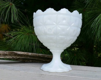 May include: A white, vintage-style glass bowl with a pedestal base. The bowl has a scalloped rim and a diamond-patterned exterior. The bowl is made of opaque glass and is set against a backdrop of green foliage.
