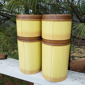 May include: Four cylindrical canisters with a pale yellow finish that fades into a darker tan at the base. Each canister features a dark brown, textured wooden lid with a visible grain pattern. The containers are arranged in two stacks of two.