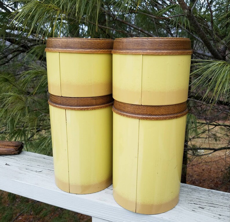 May include: Four cylindrical canisters with a pale yellow finish that fades into a darker tan at the base. Each canister features a dark brown, textured wooden lid with a visible grain pattern. The containers are arranged in two stacks of two.