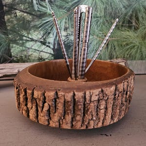 May include: A rustic wooden nut bowl with a bark-like exterior. It holds a set of silver nutcrackers and picks. The bowl is round and sits on a gray surface, with a blurred green background.