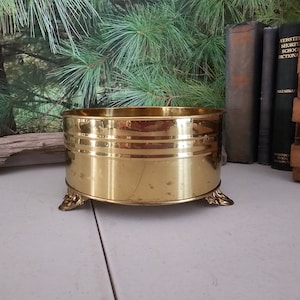 May include: A gold-toned metal planter with three decorative feet. The planter is round and has a smooth, shiny surface.