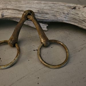 May include: A weathered metal horse bit featuring two circular rings connected by a jointed mouthpiece. The surface of the metal shows signs of oxidation and age, with a brownish, textured patina covering the entire structure of the bit.