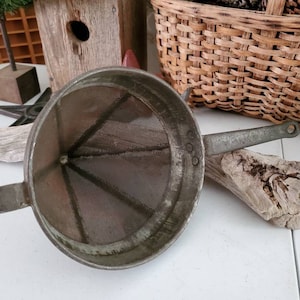 May include: A vintage metal strainer with a cone-shaped mesh and a handle. The strainer is made of metal and has a green patina. The strainer is sitting on a white surface.