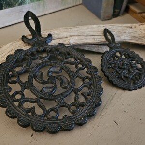 May include: Two black cast iron trivets with intricate, openwork designs. One is round, the other oval, both with a loop for hanging. The trivets are displayed on a light-colored surface with a piece of driftwood.