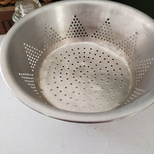 May include: A silver metal colander with a star-shaped pattern of holes. The colander is empty and sitting on a white surface.