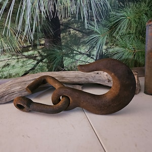 May include: A large, rusty metal hook with a curved end and a connecting loop. The hook is a dark brown color and appears to be made of heavy-duty metal. The hook is lying on a white surface.