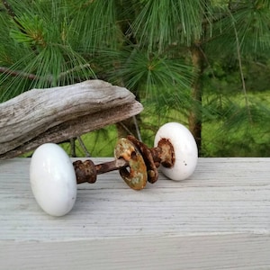 May include: A pair of white porcelain door knobs with a rusty metal connecting rod. The knobs are on a weathered wooden surface.