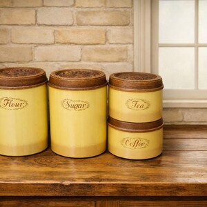 May include: A set of four cylindrical kitchen canisters in a pale yellow finish with dark brown textured lids. The canisters are labeled with cursive text reading Flour, Sugar, Tea, and Coffee, each enclosed in an oval decorative border.
