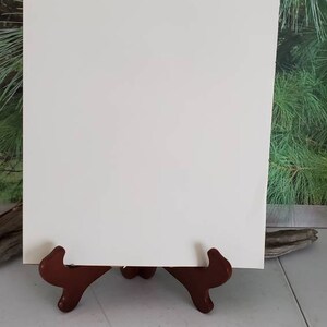 K&ouml;nnte beinhalten: Ein leeres, rechteckiges Schild aus wei&szlig;em Karton steht aufrecht auf einem dunkelbraunen, geschwungenen Holzst&auml;nder. Das Schild hat eine glatte, matte Oberfl&auml;che und saubere, gerade Kanten und bietet eine neutrale Fl&auml;che f&uuml;r Text.