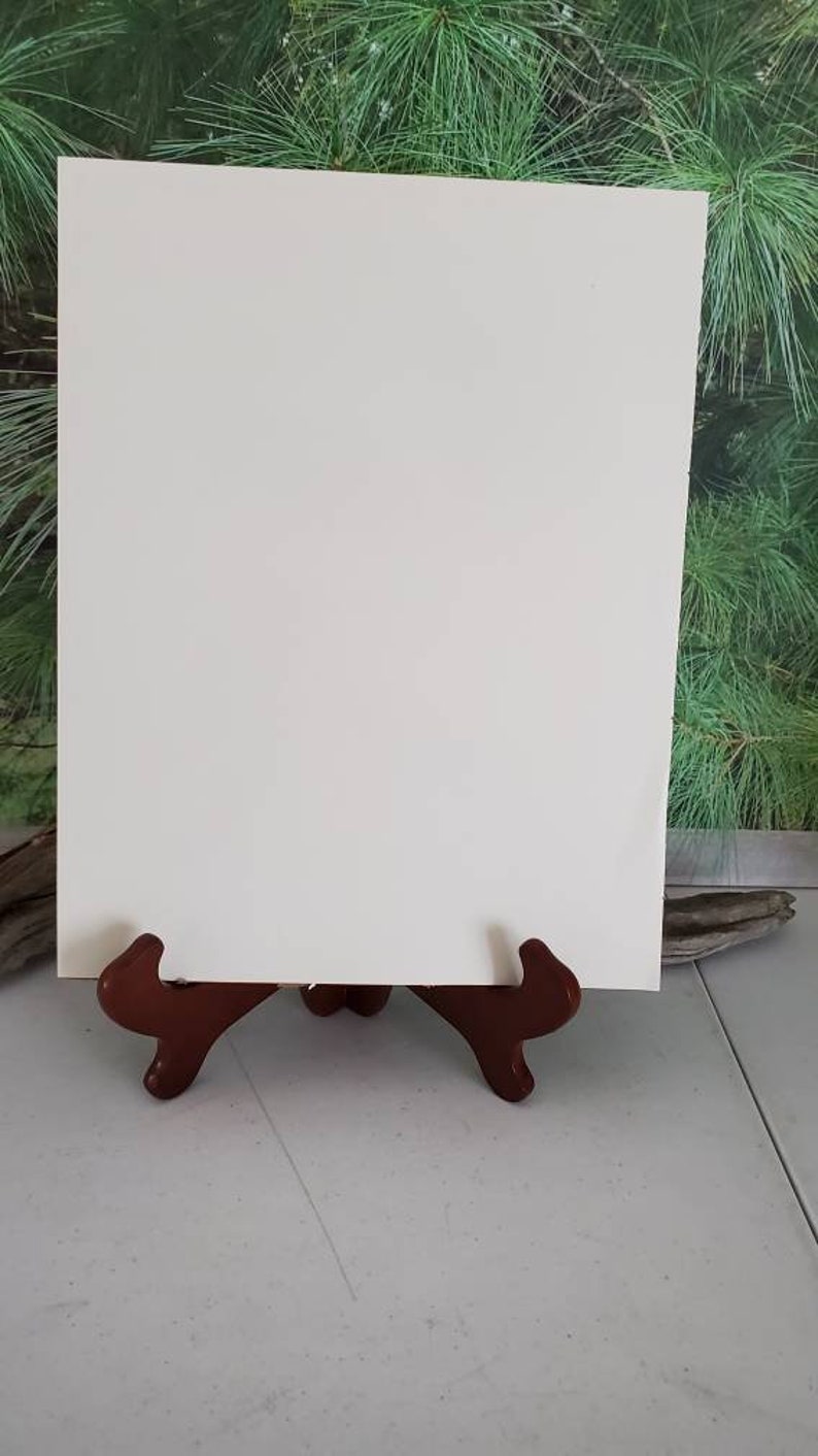 K&ouml;nnte beinhalten: Ein leeres, rechteckiges Schild aus wei&szlig;em Karton steht aufrecht auf einem dunkelbraunen, geschwungenen Holzst&auml;nder. Das Schild hat eine glatte, matte Oberfl&auml;che und saubere, gerade Kanten und bietet eine neutrale Fl&auml;che f&uuml;r Text.