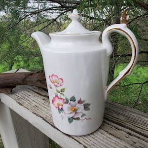 May include: White ceramic teapot with a gold trim and floral design. The teapot has a lid and a handle. The flowers are pink and white with green leaves.