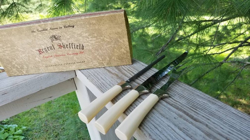 Puede incluir: Un juego de cuatro cuchillos con mangos de marfil y hojas negras en su caja original. La caja es dorada con el texto "The Renowned Name in Cutlery Regal Sheffield English Platinum Carving Set Guaranteed Forever Sharp".