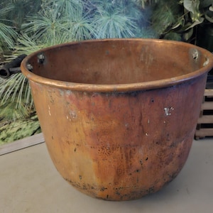 May include: A large, antique copper cauldron with a wide, rounded body and a slightly flared rim. The cauldron has a rich, reddish-brown patina and two black handles. It is likely used for cooking or decorative purposes.