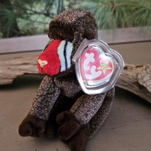 May include: A plush toy of a mandrill monkey with a red and blue face, brown fur, and dark brown limbs. The toy has a heart-shaped tag attached. The background is blurred, with green foliage visible.