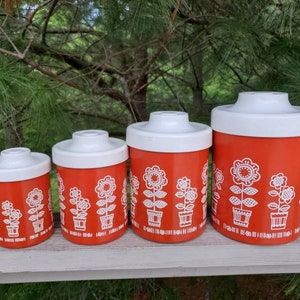 May include: Set of four red and white ceramic canisters with a floral design. The canisters are different sizes and have white lids.