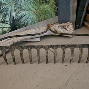 May include: An antique garden rake with a curved handle and multiple tines. The metal rake shows signs of age and wear, with a rustic, weathered appearance. The rake is a gardening tool.