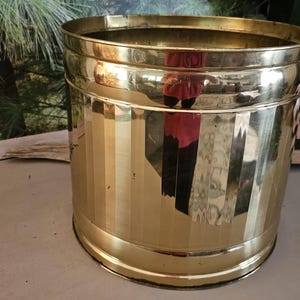 May include: A cylindrical brass wastebasket featuring a polished, reflective surface with vertical fluted detailing around the exterior. The container has a smooth, circular rim and a solid base, showcasing a metallic gold tone throughout the body.