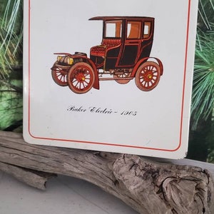 May include: A vintage white and red enamel sign featuring a black Baker Electric car from 1905. The sign has a red border and the text "Baker Electric - 1905" is printed in black.