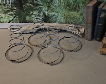May include: Four vintage metal bed springs with a spiral design. The springs are a faded silver color with a circular base. The background includes a wooden beam, books, and a wooden crate.