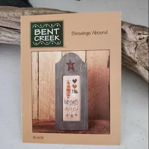 May include: A decorative wooden frame with a cross-stitch design. The cross-stitch features a pumpkin, hearts, and the words "blessings abound." The frame is on a tan background with the words "Bent Creek" and "Blessings Abound" printed on the top.