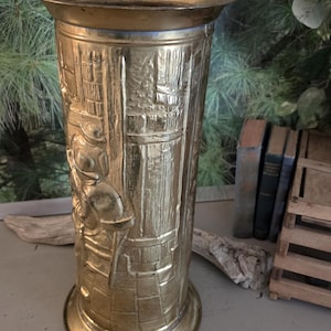 May include: A tall, cylindrical brass umbrella stand featuring a detailed embossed relief scene of a person standing in a doorway. The metallic vessel has a wide, flared rim and a circular base, showcasing a polished gold-toned finish.
