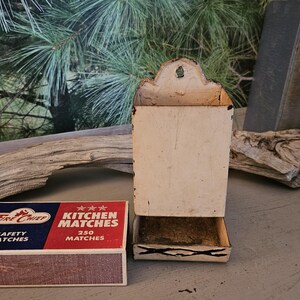 May include: A rectangular Fire Chief Kitchen Matches box with red, white, and blue branding next to a cream-colored metal wall-mounted match holder. The matchbox features the text Fire Chief, Safety Matches, and Kitchen Matches 250 Matches.