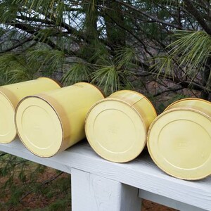 May include: A set of four cylindrical canisters in a pale yellow finish, arranged in a row. Each canister features a smooth, solid-colored exterior with a slightly darker yellow rim at the top edge, showcasing a simple, functional design.