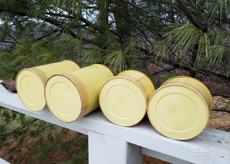 May include: A set of four cylindrical canisters in a pale yellow finish, arranged in a row. Each canister features a smooth, solid-colored exterior with a slightly darker yellow rim at the top edge, showcasing a simple, functional design.