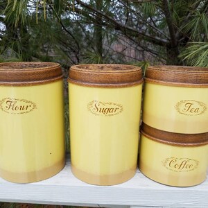 May include: A set of four cylindrical kitchen canisters with a pale yellow finish and dark brown wood-grain textured lids. The canisters are labeled with brown cursive text reading Flour, Sugar, Tea, and Coffee, each enclosed in an oval border.