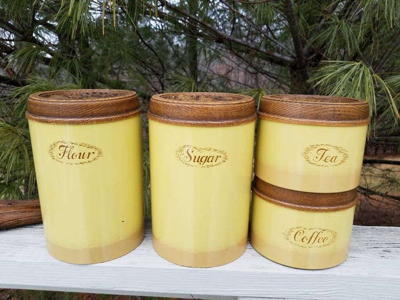 May include: A set of four cylindrical kitchen canisters with a pale yellow finish and dark brown wood-grain textured lids. The canisters are labeled with brown cursive text reading Flour, Sugar, Tea, and Coffee, each enclosed in an oval border.