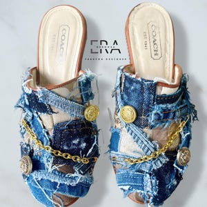 May include: A pair of Coach mules featuring a patchwork design of blue denim fabric with frayed edges, layered over a tan monogram print. Each shoe is accented with two gold-toned metal buttons and a gold chain draped across the front upper.