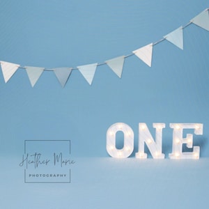 Könnte beinhalten: Ein hellblauer Hintergrund mit einem Banner aus dreieckigen Flaggen und einem Leuchtschild "ONE". Das Schild ist weiß mit kleinen Lichtern im Inneren. Das Bild enthält auch den Text "Heather Marie Photography".