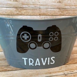 May include: A gray metal storage bin with a black and white video game controller graphic and the name "TRAVIS" in white lettering.
